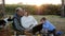Happy mature couple on picnic with grandson and pet sits at plaid with laptop on background sunrise