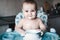 Happy messy baby boy sitting in highchair and playing with his dirty plate from yogurt