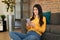 Happy mexican lady using smartphone, chatting with friend or lover, sitting on floor by sofa at home, copy space