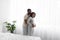 Happy millennial african american man in home clothes, hugs pregnant wife in white bedroom interior on window background