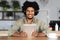 Happy millennial black curly guy in headphones looking at tablet, surfing in internet, sitting at sits at table with cup