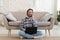 Happy millennial european guy with stubble in shirt calls by phone, works on laptop, sits on floor in living room