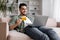 Happy millennial islamic guy with beard in casual sits on sofa with laptop show credit card in living room interior