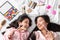 Happy millennial korean woman and teen girl, hold brushes, lie on bed with cosmetics in bedroom, top view