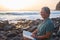 Happy moment for a senior woman alone sitting on the beach of pebbles reading a book. 70 years old. Waves of the ocean. Sunset