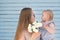 A happy mother and daughter toddler are sniffing a small bouquet of white flowers against a blue planked wall. Scene of love,