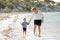 Happy mother holding hand of sweet blond little daughter walking together on sand at beach sea shore