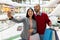 Happy multinational couple holding gift bags, taking selfie at supermarket, going shopping together