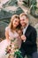 Happy newlyweds look on a photographer. Man and woman in festive clothes sit on the stones near the wedding decoration