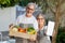 Happy old european wife, husband in aprons hold box with organic vegetables, show smartphone with blank screen