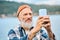 Happy older hipster man standing in nature park taking photos on smartphone.