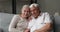 Happy older spouses sit on couch hug look at camera