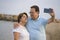 Happy pensioner woman and her husband taking romantic walk taking selfie - happy retired mature couple walking on the beach during