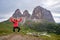 Happy photographer jumping with her arms up in the air, wearing a bright red jacket, in front of Sassolungo Saslonch, Langkofel