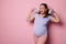 Happy pregnant woman holds blue knitted baby booties, smiles broadly looking at camera, isolated on pink background.