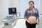 Happy pregnant woman standing in consulting room after ultrasonography near ultrasound scan with fetus 3d image on monitor screen