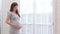 Happy Pregnant Woman standing in front of windows and stroking her big belly with love at cozy home,Pregnancy of young woman enjoy