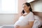 Happy pregnant woman talking on mobile phone, sitting alone in a light bedroom interior. Home relax, pregnancy concept