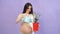 Happy pregnant woman in underwear watering potted plant over purple background.