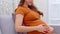 Happy Pregnant Woman wear brown dress sitting on couch holding stroking her big belly at home,Pregnancy young woman enjoying with