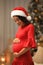 Happy pregnant woman wearing Santa hat in room for Christmas. Expecting baby