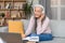 Happy pretty retired old european gray-haired lady in wireless headphones working with laptop in living room