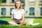 Happy pretty student woman is sitting on the lawn in the university campus with notebook in her hands and useing laptop.