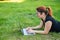 Happy red-haired girl in glasses lies on the lawn in the park and types on the laptop keyboard. Young caucasian female