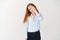 Happy redhead woman smiling with half face covered behind hand, looking carefree at camera, standing over white