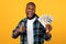 Happy Rich Black Guy Holding Money Gesturing Thumbs-Up, Yellow Background