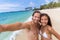 Happy seflie couple taking picture with smartphone on beach vacation smiling at camera. Asian woman, Caucasian man