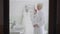 Happy senior Caucasian bride in bathrobe standing in bathroom admiring reflection in mirror and wedding dress hanging at