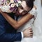 Happy sensual handsome groom hugging beautiful bride with bouquet in city streets closeup
