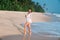 A happy, shining young girl walks along the beach on the island, holds a delicious cold coconut in her hand, smiles and