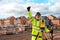 Happy Site engineer in hi-viz working on house building construction site using modern surveying equipment against new houses nad
