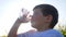 Happy small guy drink clean water on nature on background field flowers, little boy drinking from glass outdoors,child