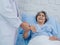 Happy smile beautiful Asian elderly old woman patient in light blue dress lying on bed while male doctor in white suit hold hand.