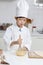 Happy smiling Asian boy in white chef uniform with hat at kitchen, stirring and mixing eggs in glass bowl, little cute baker child