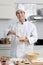 Happy smiling Asian man in white chef uniform with hat, stirring and mixing eggs in glass bowl. Portrait of young mam cooking meal