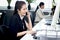 Happy smiling Asian woman officer sitting, talking on mobile phone at office desk with blurred busy colleague background, modern