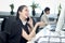 Happy smiling Asian woman officer sitting, talking on mobile phone at office desk with blurred busy colleague background, modern