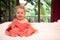 Happy smiling baby in soft pink blanket on white bed sheets in villa bedroom looking at camera with blurred tropical background an