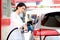 Happy smiling customer woman standing and waiting for filling up her car with fuel petrol pump nozzle against, beautiful young