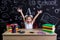 Happy, smiling and excited schoolgirl sitting at the desk with both arms up, surrounded with school supplies. Chalkboard