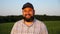 Happy smiling farmer in cap looks into camera standing at cornfield. Portrait of adult agronomist with beard with green