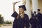 Happy smiling graduate girl dressed in black gown raises up her hand holding a diploma.