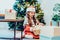 A happy smiling little girl in Santa hat and mom`s festive knitted sweater packing gift boxes on the Christmas tree background.