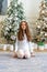 Happy smiling little girl sitting on the carpet by the Christmas tree at home waiting for gifts in the morning