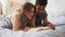 Happy smiling multiethnic couple lying in bed reading a book together at home in bedroom. Slowmotion shot