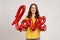 Happy smiling romantic teenager girl in yellow casual T-shirt holding foil balloon letters, looking at camera, expressing love and